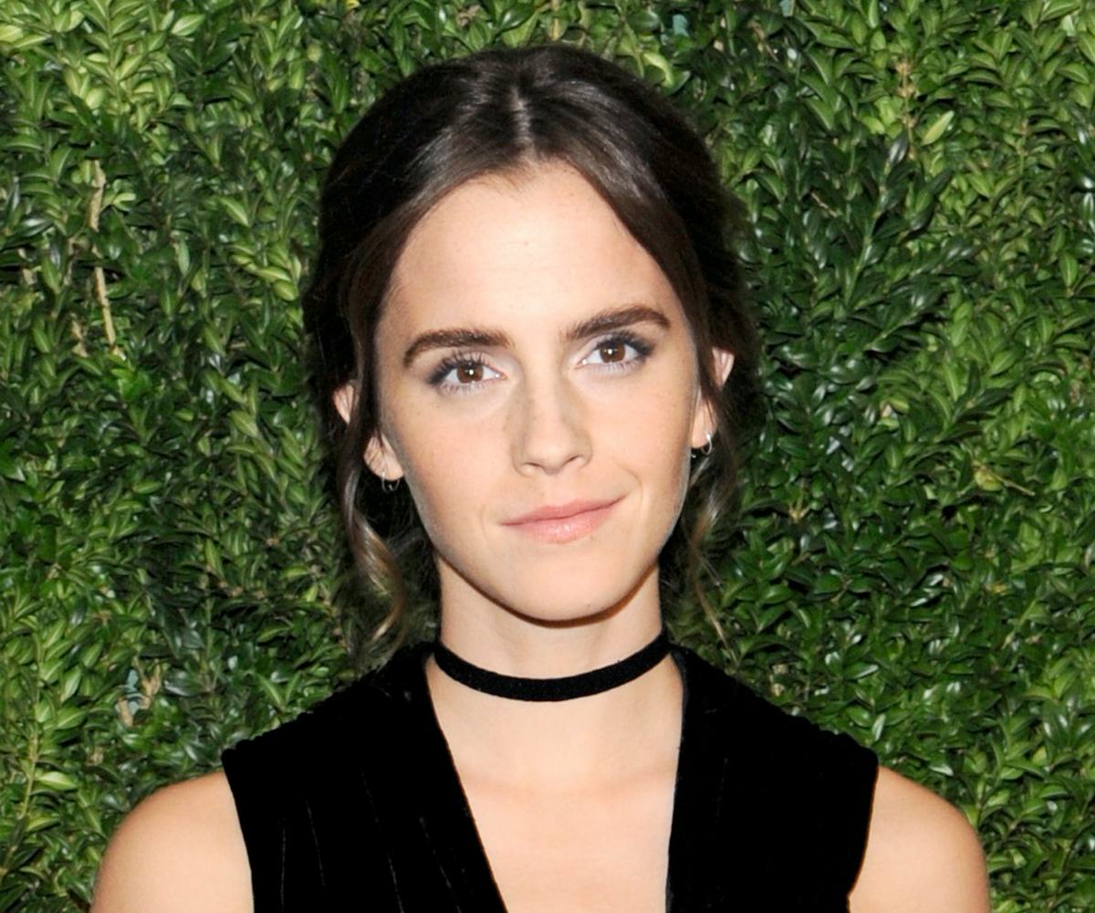 Emma Watson smiling while wearing a black dress and a black velvet choker in front of a green backgr...