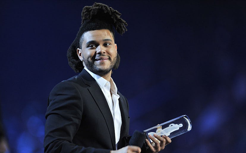 The Weekend wearing a black suit and white shirt while holding a mic in one hand and an award in the...