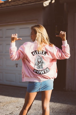 A blonde woman pointing at her back in a pink jacket from NYLONshop and Petals and Peacocks collabor...