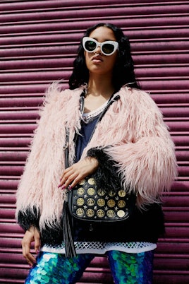 A model in a baby pink furry coat and white sunglasses carrying a black coin shoulder bag by Poppy L...