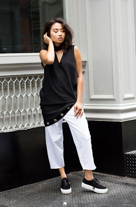A girl with brown hair posing in a black top and white pants with black and white shoes