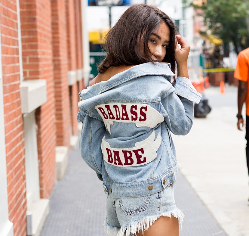 A girl walking down the street in a white top, denim shorts, and a denim jacket with "Badass babe" t...