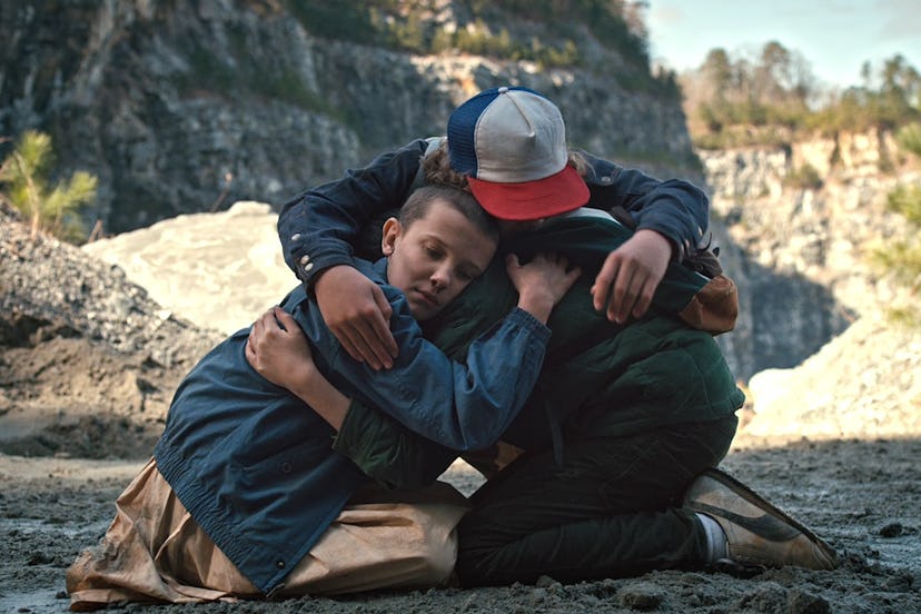 Eleven, Mike, and Dustin are hugging each other on the ground during the sunny day in the Stranger T...