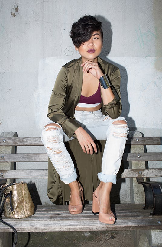 Lynn Kim wearing light ripped jeans, a burgundy top, and an olive jacket while sitting on a bench 