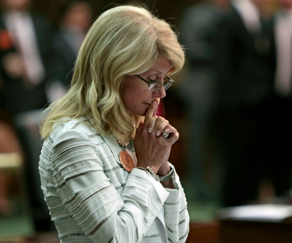 Wendy Davis wearing a white striped blazer, a pearl necklace, and glasses while looking down pensive...