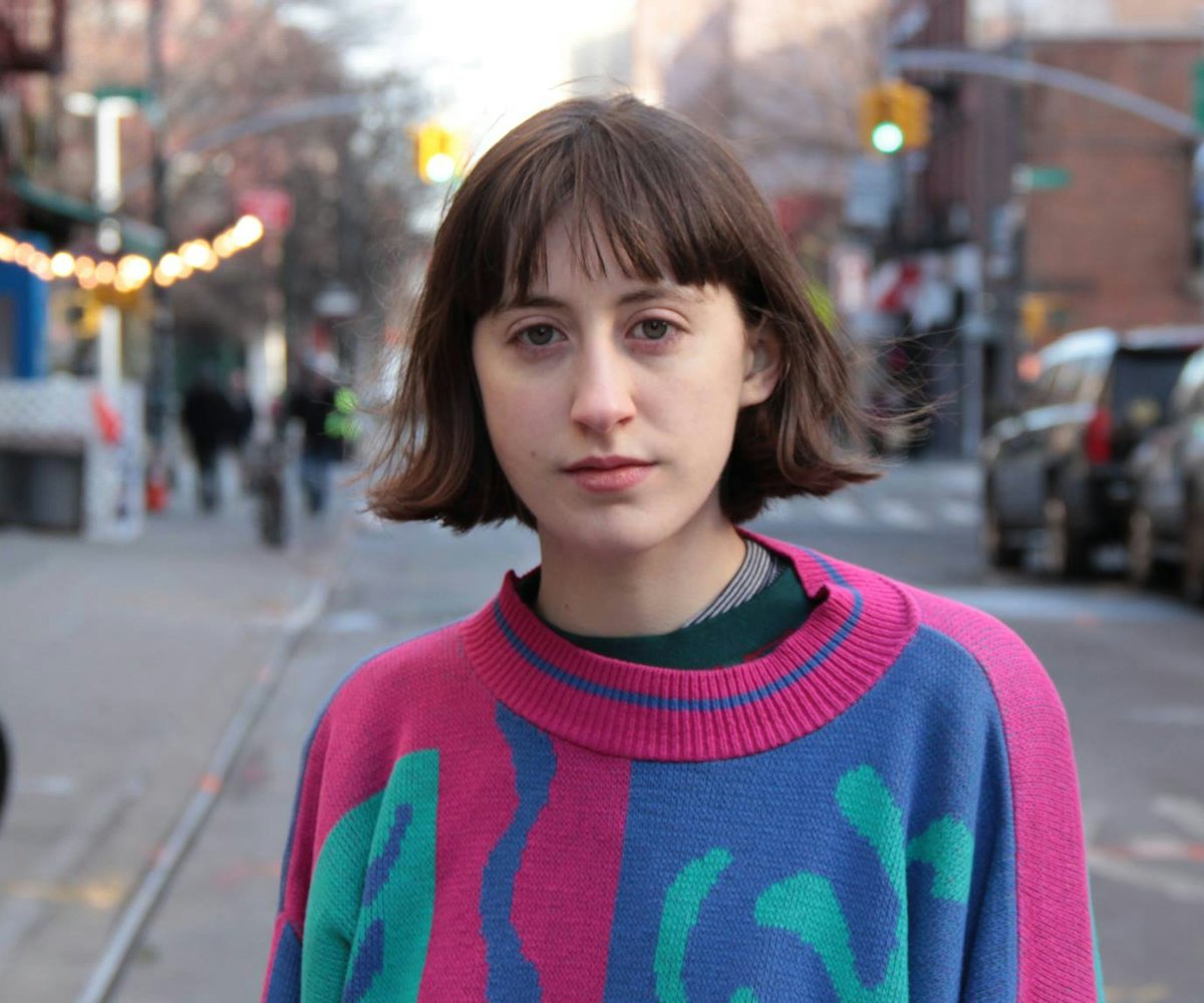 Frankie Cosmos (Greta Kline) standing in the street wearing a sweater with pink, blue and green patt...