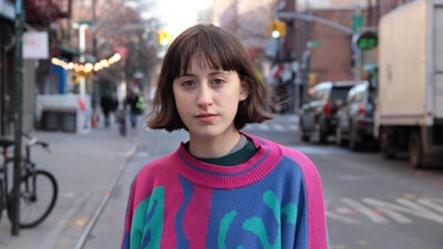 Frankie Cosmos (Greta Kline) standing in the street wearing a sweater with pink, blue and green patt...