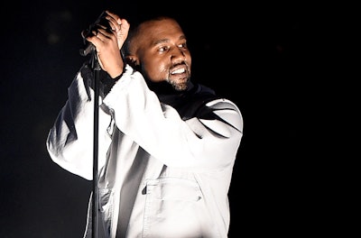 Kanye West on stage in an all white outfit singing and smiling as the spotlight is directly on him