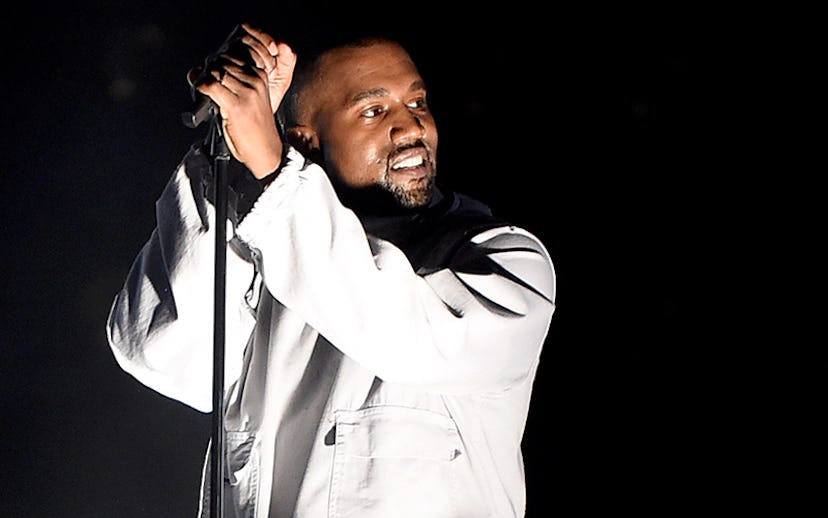 Kanye West on stage in an all white outfit singing and smiling as the spotlight is directly on him