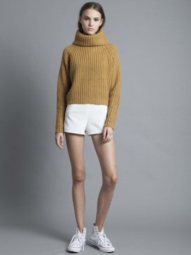 Woman with brown hair wearing a beige Lioness, Camel turtleneck knit, white shorts, and white sneake...
