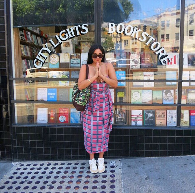 A black-haired woman posing in front of a "City Lights Bookstore" while wearing a purple dress