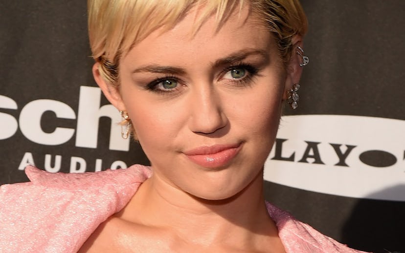 Miley Cyrus posing on a red carpet in a beige top and pink jacket 