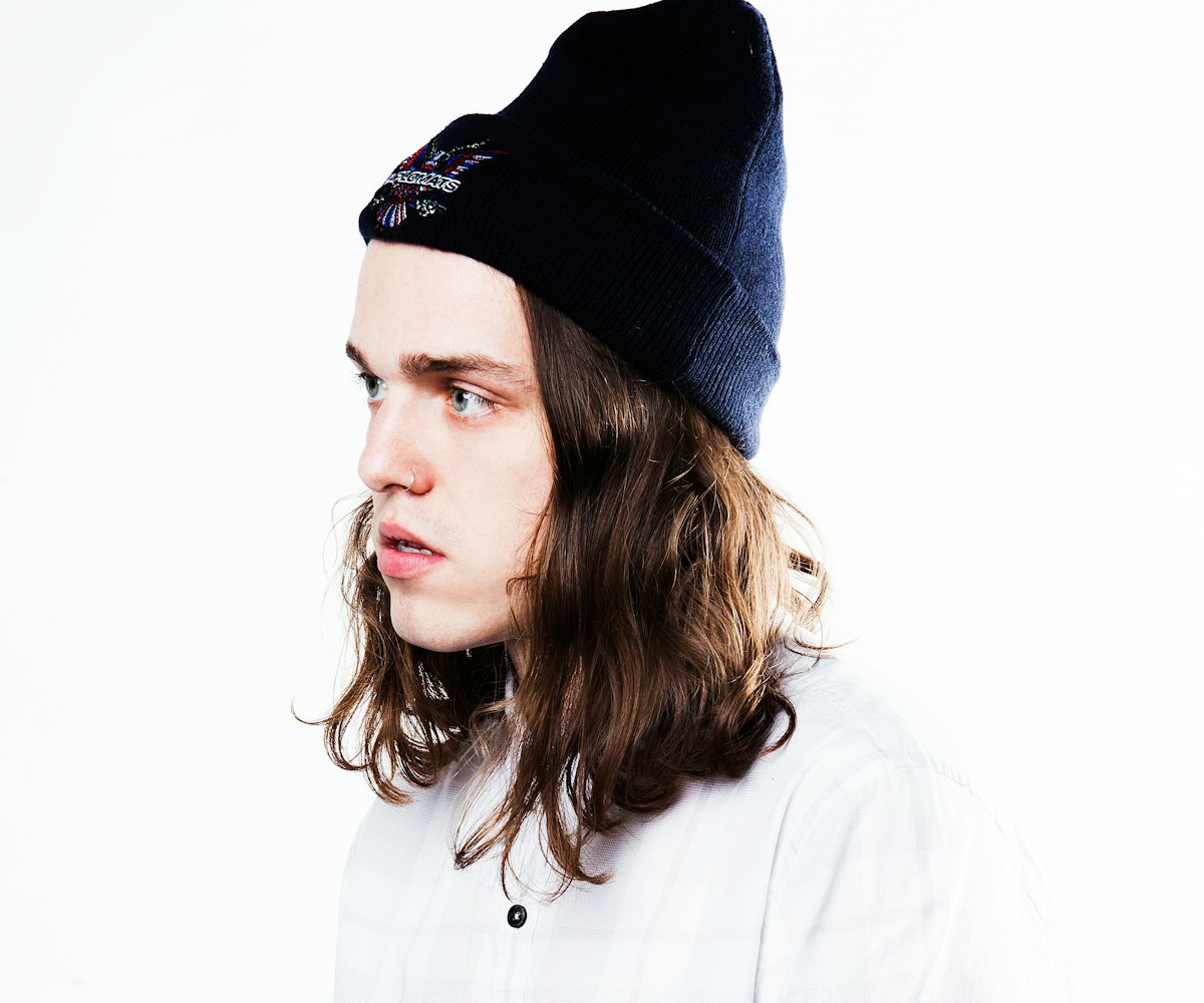 A male model posing in a white shirt and a black beanie with mid length brown wavy hair
