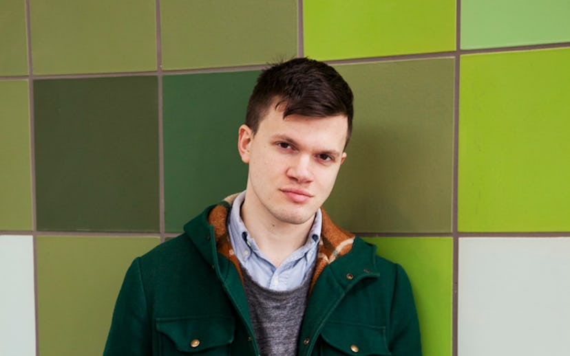 Sasha Chapin in a light-blue shirt, grey sweater, and dark green jacket in front of a green-orange-b...