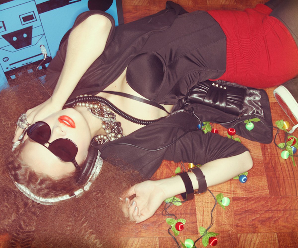 A woman lying on the floor in a black satin corset, black jacket, and red shorts with large headphon...
