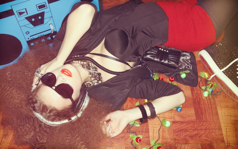 A woman lying on the floor in a black satin corset, black jacket, and red shorts with large headphon...