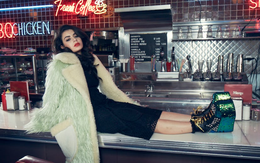 Charlie XCX wearing a mint fur coat, a black dress, and YRU platform boots while sitting on a counte...