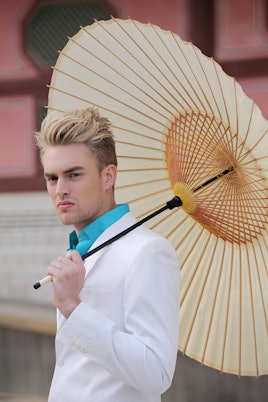 A young man posing in a light blue shirt and a white sweater while holding a brown umbrella