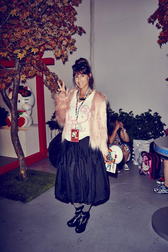 A lady smiling and wearing a Hello Kitty white tank top with a pink feather coat and wide black sati...