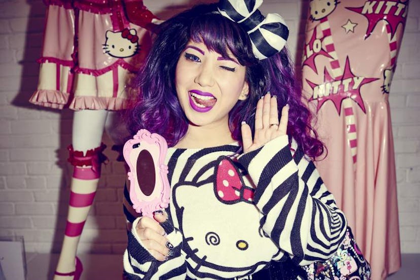 A  young girl with purple hair holding a mirror in her hand and winking in a Hello Kitty striped shi...