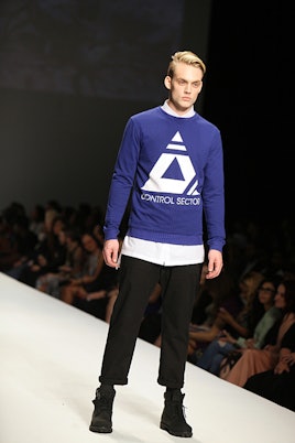 Top Model contestant walking down a runway while wearing black pants combined with a blue sweater ov...