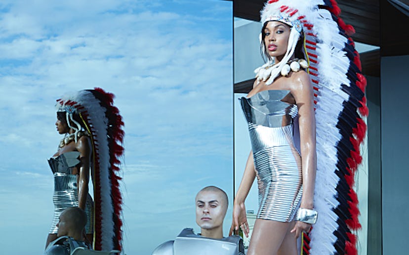Black woman in futuristic outfit and silver high heels wearing war bonnet while standing next to rob...