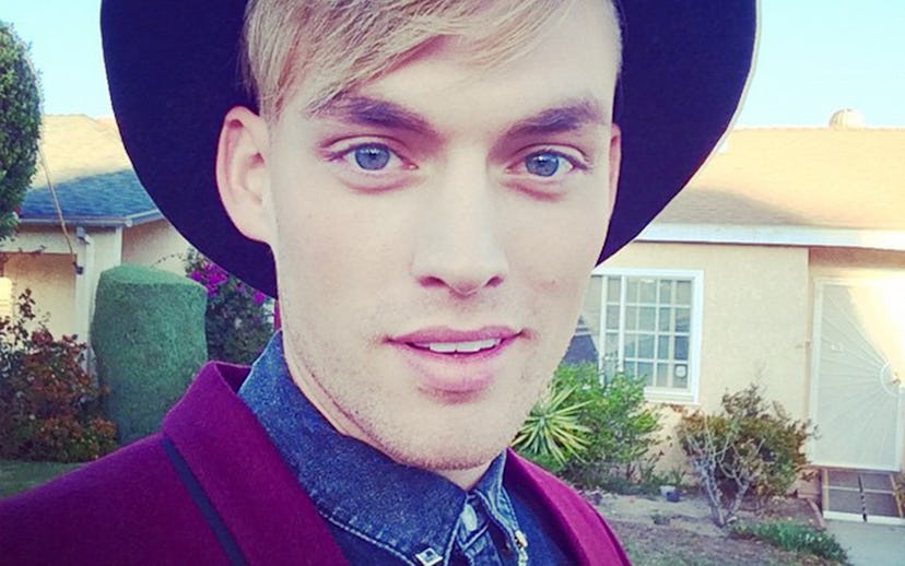 A man with blonde hair swept to the side wearing a round hat, a jean shirt with chains on the collar...