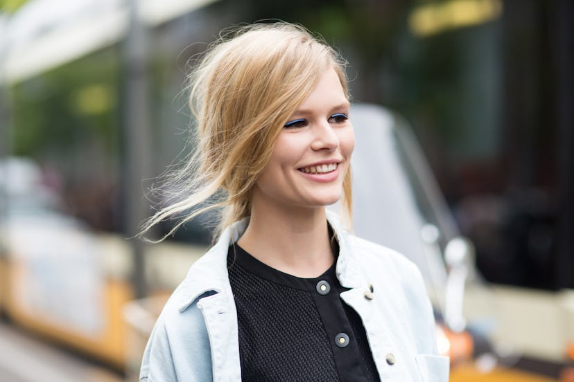 A blonde woman smiling while wearing a black shirt and light blue coat with fair blue eyeshadow