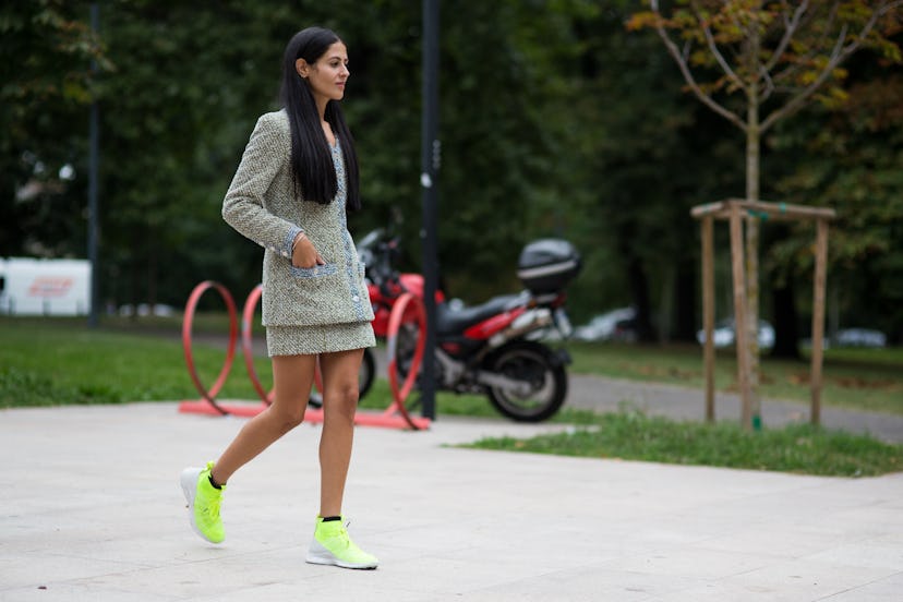 A brunette woman in a grey plaid long blazer and matching skirt, and lime-green and white sneakers w...