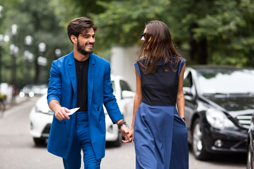 A man in a blue suit and black shirt smiling holding hands with a woman in a black sleeveless shirt ...