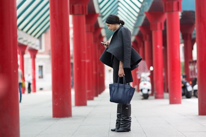 A brunette woman in a black jacket and black knee-high boots, black dress and dark navy bag