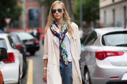 A blonde woman posing in a light brown coat, white shirt, and a black scarf with floral pattern