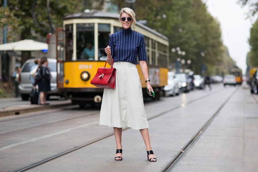 A woman in a dark blue shirt with white stripes, a cream midi skirt, red purse and black sandals