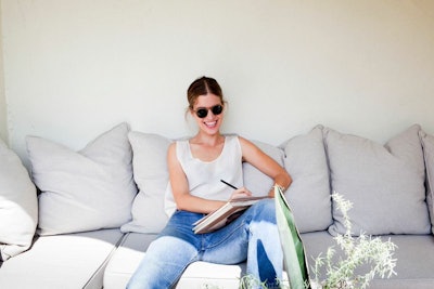 A girl in sunglasses, a white tank top and jeans sitting on a grey couch and writing a letter using ...