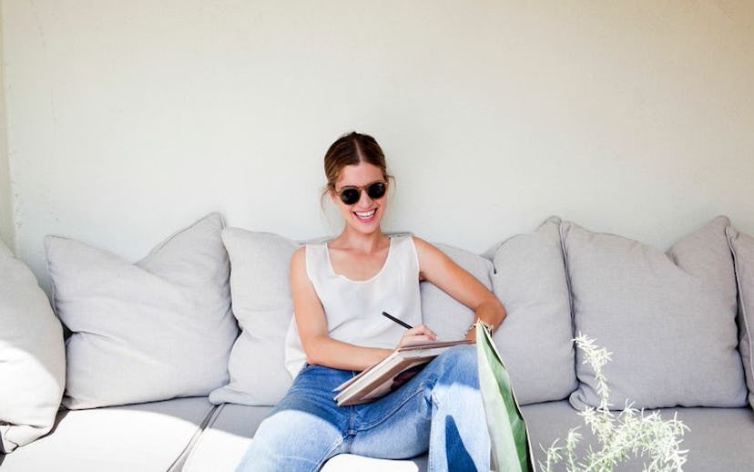 A girl wearing a white tank top and jeans while sitting on a grey couch and writing a letter while u...