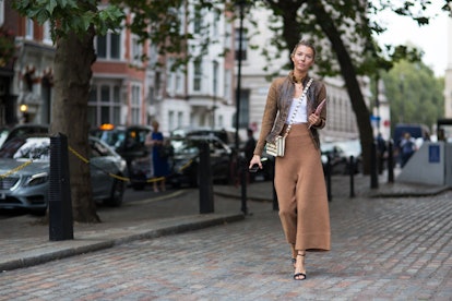 A woman wearing brown wide-leg pants, a brown leather jacket, a striped purse, and black sandals whi...