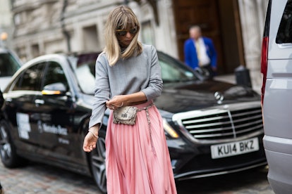 A woman with sunglasses on, a pink skirt, a grey sweater, and a purse with snake-skin print