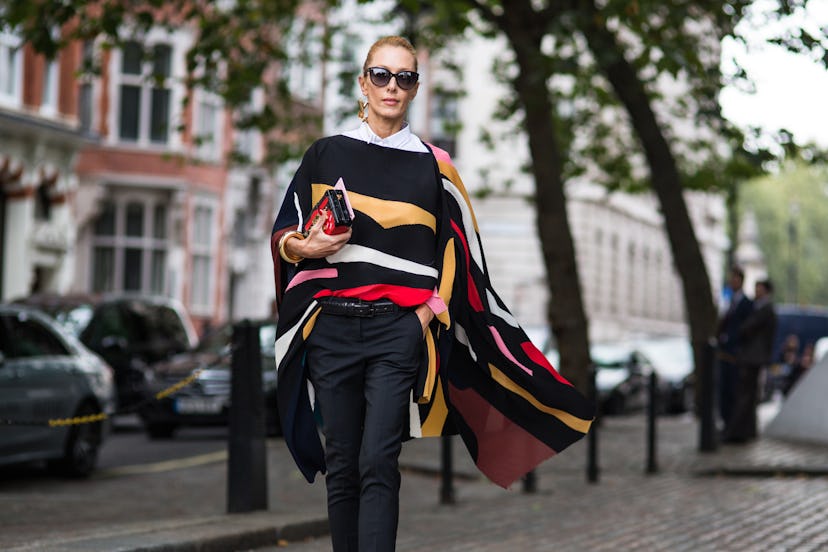 Elina Halimi with sunglasses on, wearing a colorful poncho sweater, black pants, and a red & black p...