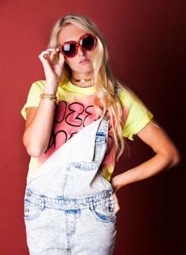 Blonde woman with heart-shaped glasses, a yellow shirt, and denim overalls