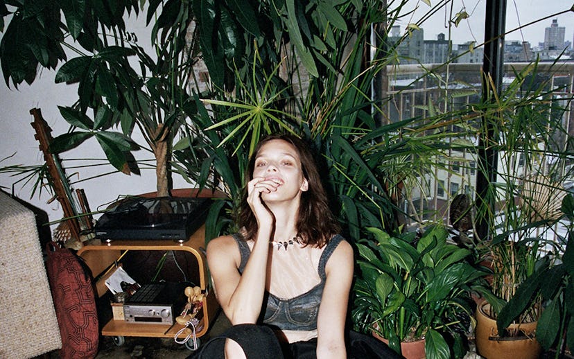 A girl posing in front of home plants in a tulle-denim top, black skirt, and a choker with spikes