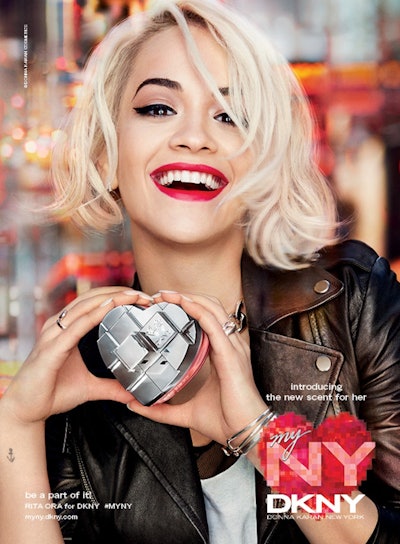 Rita Ora, with her blonde hair and red lipstick, smiles while holding a heart shaped MYNY scent by D...