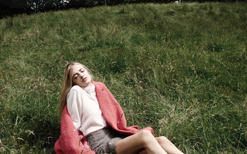 A girl wearing a pink coat, a white turtleneck, and a plaid mini skirt sitting on the grass