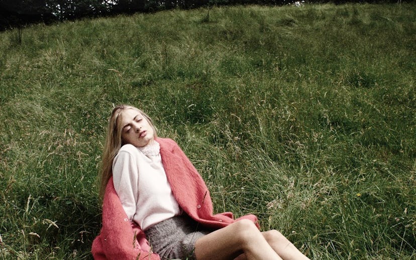 A girl wearing a pink coat, a white turtleneck, and a plaid mini skirt sitting on the grass 