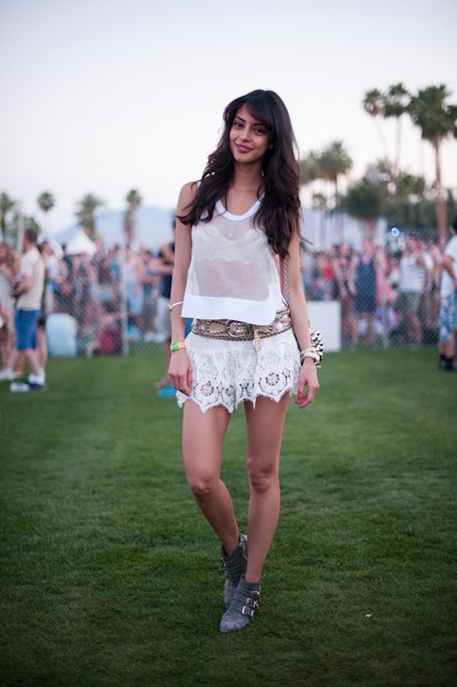 A woman with long dark hair wearing a translucent tank top, short white lace skirt, and beige belt