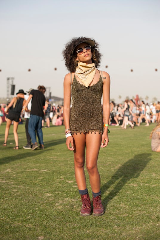 A woman in a short strappy olive-black dotted dress, beige bandana around her neck, burgundy boots a...