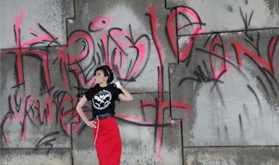 Kristeen Young standing in front of a graffiti-painted wall in a red skirt, black t-shirt with a whi...