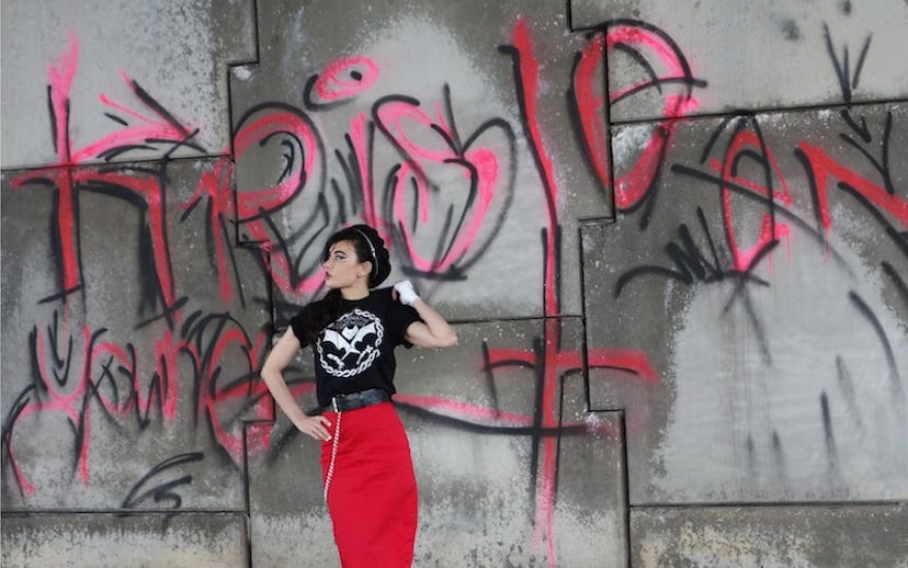 Kristeen Young standing in front of a graffiti-painted wall in a red skirt, black t-shirt with a whi...