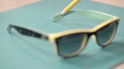 Yellow Ray-Ban sunglasses lying on a blue surface with a pen pointing to an engraving "Rock"