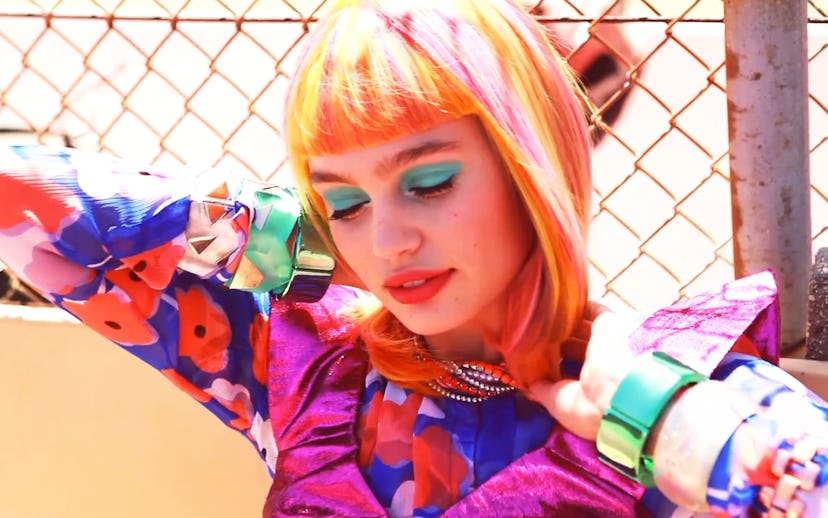 A model with orange and pink hair, turquoise eyeshadow, and peach lipstick posing in a colorful outf...