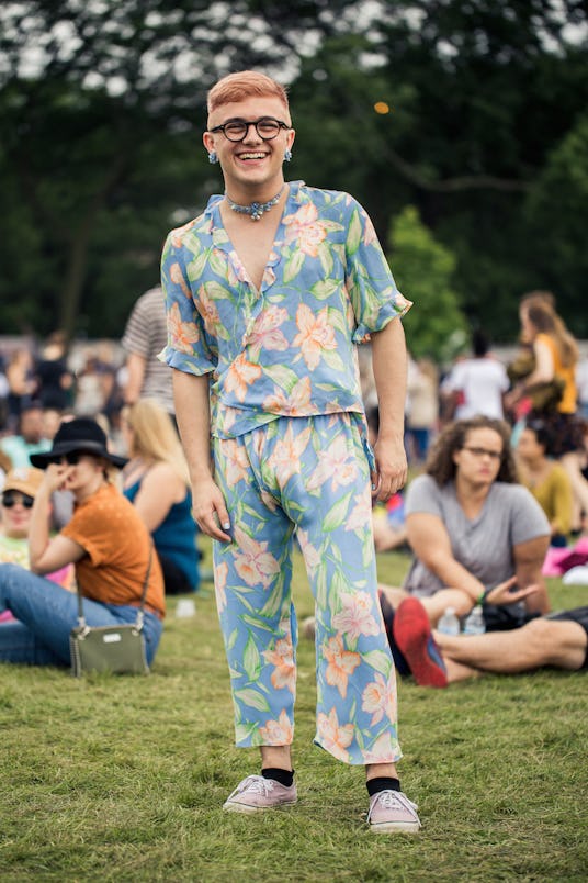 A man with pink hair, circle glasses, blue earrings and necklace, and a blue floral set of pants and...